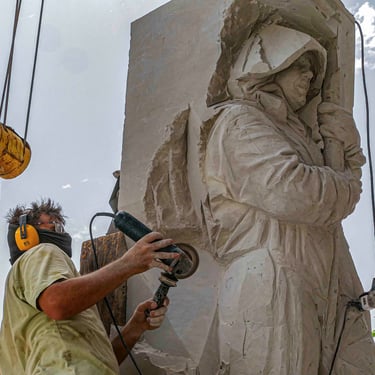 Professional sculptor using a power grinder to carve a detailed stone statue of a hooded figure.