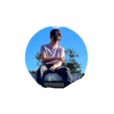 A young man in a purple t-shirt and sunglasses sitting outdoors against a clear blue sky.