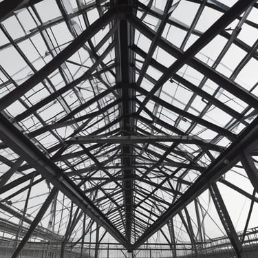 An industrial structure with a scaffolding system against a large concrete wall. The scaffolding features metal beams, platforms, and red sections, casting intricate shadows onto the wall. The concrete surface is smooth with subtle vertical lines.