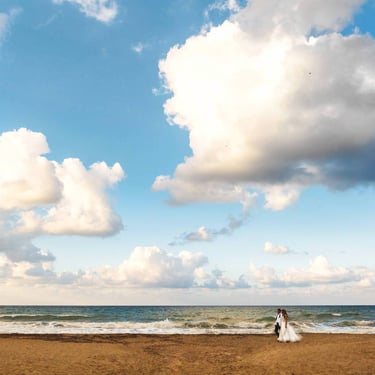 Una coppia passeggia su una spiaggia, su di loro il cielo azzurro tempestato da centinaia di nuvole 