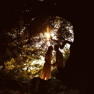 Una coppia si tiene per mano in un bosco in montagna, illuminati dai raggi solari filtrati dai rami di un albero secolare