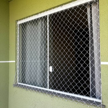 janela de correr de alumínio em parede verde com rede de proteção instalada externamente; luz do sol incidindo e fixações vis