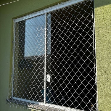 janela de correr de alumínio em parede verde com rede de proteção instalada externamente; luz do sol incidindo e fixações vis