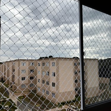 janela ou sacada vista por dentro com rede de proteção; moldura de alumínio dividindo a abertura e, ao fundo, prédios residen
