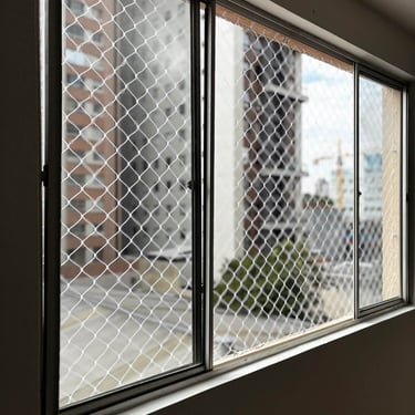 janela de correr de alumínio vista por dentro com rede de proteção instalada; ao fundo, prédios residenciais em dia claro
