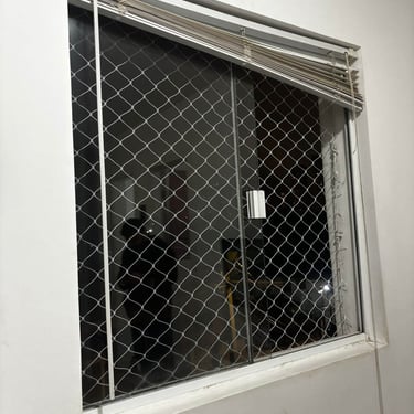 Rede de proteção fixada por dentro da janela com persiana e trilhos aparentes.