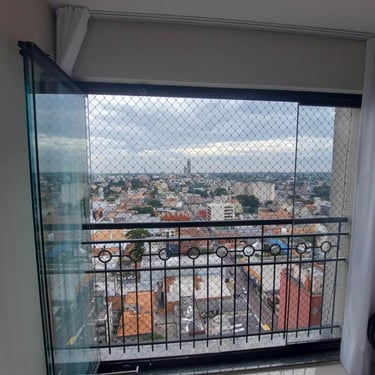 Sacada envidraçada com rede de proteção e guarda-corpo de ferro; vista ampla da cidade em dia nublado.