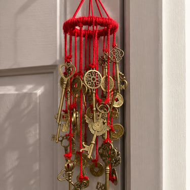 Handmade windchime in red yarn with hanging brass keys and Chinese cash coins, photographed indoors against a door
