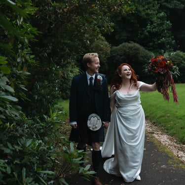 A bride in a white gown and a groom in a Scottish kilt walk through a lush garden path.