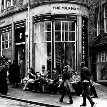 Several people sit outside the Milkman, on a nice day,as others pass them on Cockburn Street, Edinburgh