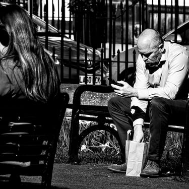 A man sit on a bench, phone in hand, digging into a paper bag for his lunch. Gardener's Crescent