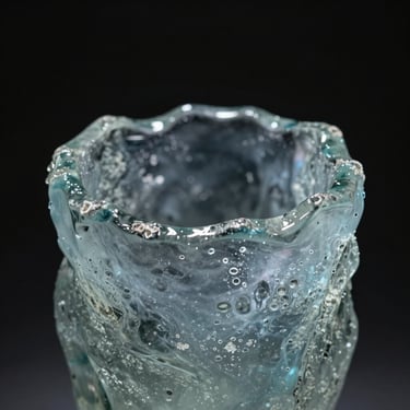 A close-up of a heavy, hand-cast glass sculpture with raw, textured edges, catching a single beam of cold slate-blue light, dark mysterious background.