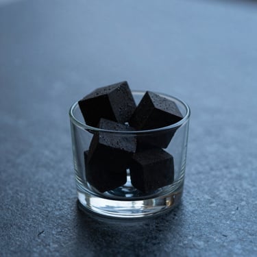 A dark, minimalist glass container holding solid scent blocks, resting on a polished dark stone surface under soft, diffused blue-hour light.