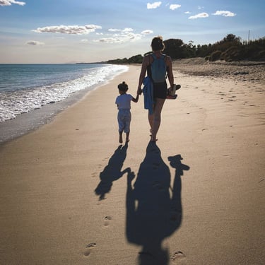 mother holds toddler's hand as they walk along a beach