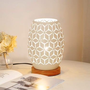 Modern white ceramic table lamp with geometric cutouts and a wooden base on a bedside table.