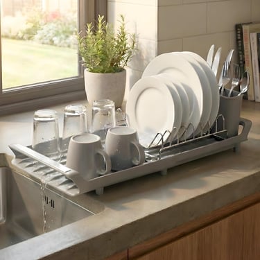 Gray plastic dish drying rack with a self-draining spout holding clean plates, mugs, and glasses on a kitchen counter.