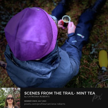 close up photo of a person wearing outdoor clothes sitting on moss holding a mug with liquid in itg