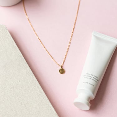Overhead flatlay of keepsake items including a delicate gold necklace, a linen journal, and a tube of hand cream on a pastel pink surface, soft daylight.