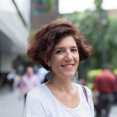 A smiling woman with short auburn curly hair wearing a white shirt and drop earrings outdoors.