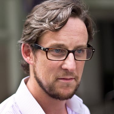 Professional man with brown hair and beard wearing tortoise shell glasses and a striped dress shirt.
