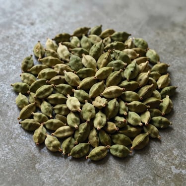 Symmetrical overhead shot of raw green cardamom pods piled on a smooth concrete floor, soft natural window light, muted organic tones.