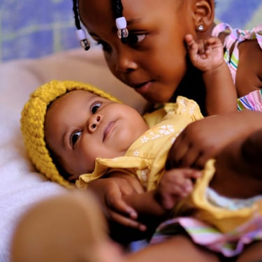 Young Black girl with braided hair gently holding her baby sister wearing a yellow knit hat.