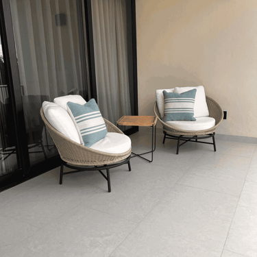 Sillas de mimbre con cojines blancos y mesa lateral en terraza con ventanal de cristal | KARÄNA
