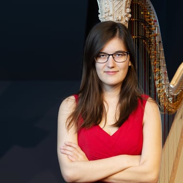 Clara Busemann steht mit verschränkten Armen vor einer Konzertharfe und schaut mit einem kleinem lächeln in die Kamera
