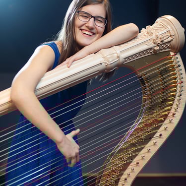 Eine Harfe liegt auf dem Boden mit der Säule nach oben. Clara Busemann hockt lachend hinter der Harfe und spielt die harfe