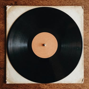 Close-up overhead shot of a worn vinyl record sleeve on a wooden surface, warm tungsten light, desaturated film-stock palette, grain texture visible
