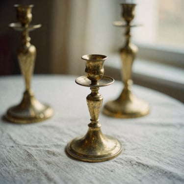 Warm editorial close-up of weathered brass candlesticks on a rumpled linen tablecloth, soft warm morning light, shallow depth of field, 35mm film