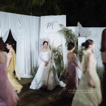 เจ้าสาวยืนกลางฉากงานแต่งโทนขาว ขณะเพื่อนเจ้าสาวเดินผ่านสร้างเอฟเฟกต์เบลอ ภาพงานแต่ง Outdoor ตอนกลางคืน