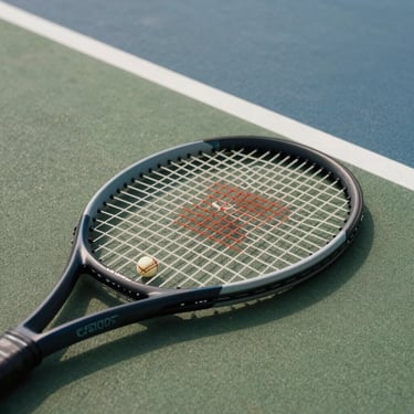 Overhead close-up of a tennis racket resting on a hard court surface, natural diffused daylight, court geometry visible in background, no people, minimal editorial framing