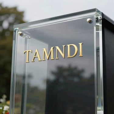 Modern transparent funeral plaque with gold lettering, reflecting soft trees under an overcast sky