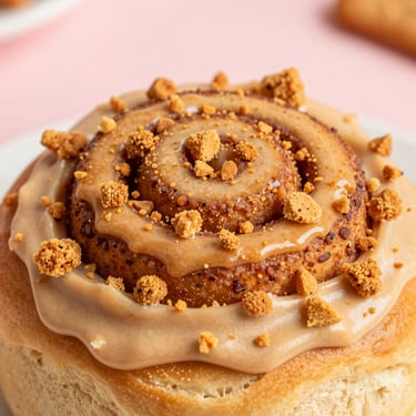 Extreme close-up of a cinnamon roll smothered in speculoos cookie butter frosting, topped with crunchy crushed Lotus biscuit pieces, warm golden tones