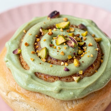 Close-up of a cinnamon roll topped with vibrant green Sicilian pistachio cream frosting and chopped toasted pistachios, bright natural light