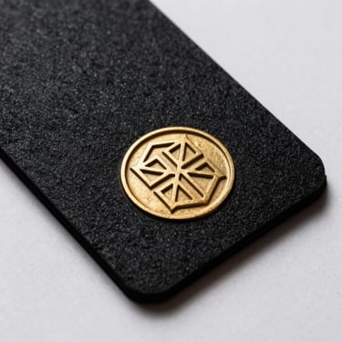 Macro photograph of a heavy-weight textured black cotton card with a gold-leaf debossed geometric seal. Minimalist composition, dramatic side light casting long shadows.