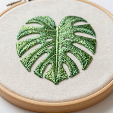 Macro overhead shot of a finished green monstera leaf embroidered on cream linen in a light wood hoop