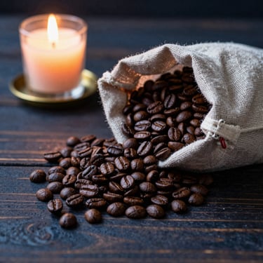 Dark roasted specialty coffee beans spilling from a rustic fabric bag onto a dark wooden table, warm incandescent light
