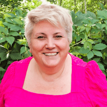 Naeratav lühikeste blondide juustega naine, kes kannab õues erkroosat toppi ja selja taga rohelisi l