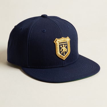 Flat-lay of a structured snapback cap in navy with gold embroidered shield icon on front, brim flat, natural side light, clean light background