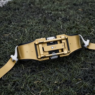 Close-up overhead shot of a youth flag football belt and flags in gold laid on a dark synthetic turf surface, natural daylight, sharp detail on the clip hardware