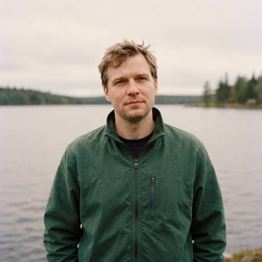 Candid portrait of a thoughtful man in his 30s outdoors near a Finnish lake, soft overcast daylight, editorial style