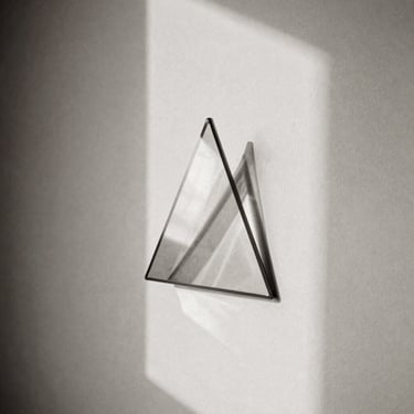 An open geometric glass prism casting complex light refractions and subtle shadows on a plain cream wall, natural soft light, black and white photography