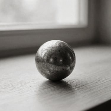 A solid aged brass sphere resting slightly tilted on a minimalist wooden surface, soft side light, deep shadows, black and white photography, sepia tint