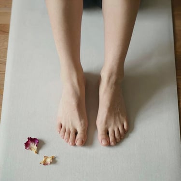 Overhead square crop of a pair of bare feet resting on a pale yoga mat, a few dried flower petals nearby, soft natural light from above, minimal and serene