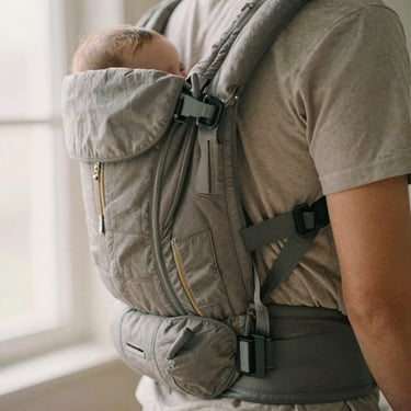 An ergonomic baby carrier in neutral gray fabric worn by a father, close-up on the secure buckles, warm domestic light