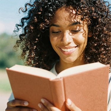 Candid medium close-up of a smiling Black woman with curly hair holding a terracotta-colored book, bright warm sunny daylight, 35mm film look