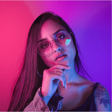 A young woman with long hair and glasses posing under vibrant pink and blue neon studio lighting.