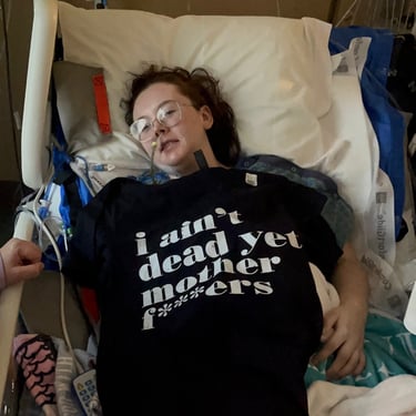 Baylie Phillips in a hospital bed with a feeding tube and shirt that reads "I ain't dead yet mother f***ers"